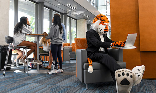 Aubie using a computer in the lounge near some students in Horton-Hardgrave Hall
