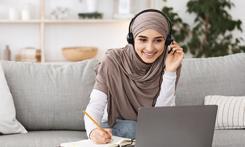 A woman wearing hijab attending and taking notes for an online class on her laptop